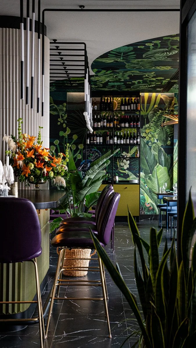 Restaurant interior with purple bar stools at pale green bar with peacock mural on wall and ceiling