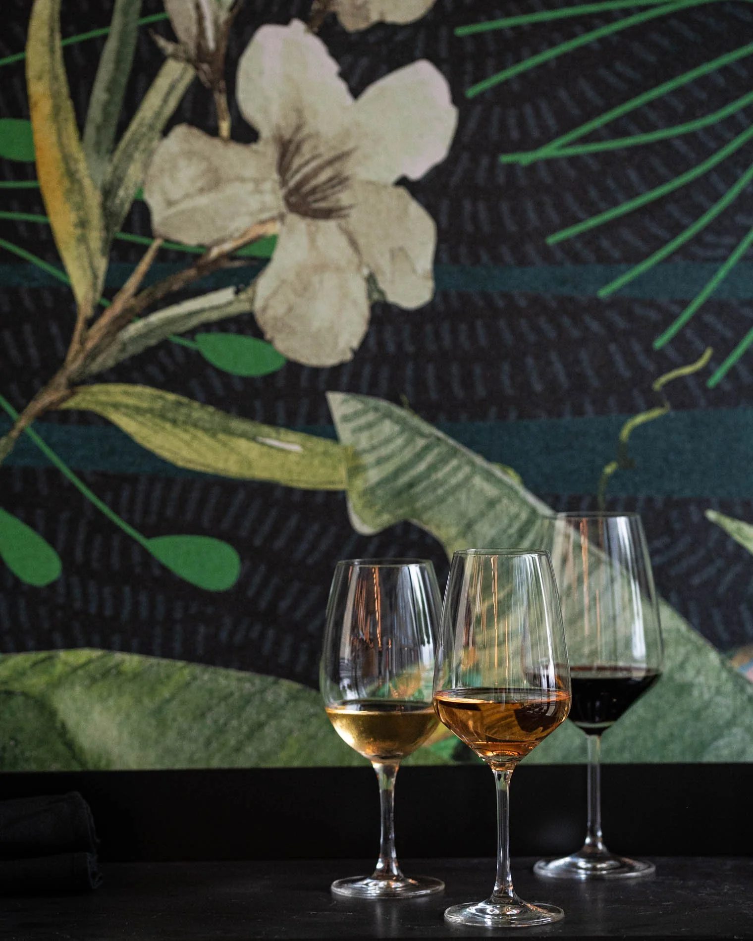 Close up of 3 wine glasses on black stone counter with floral and tropical plant wall mural behind