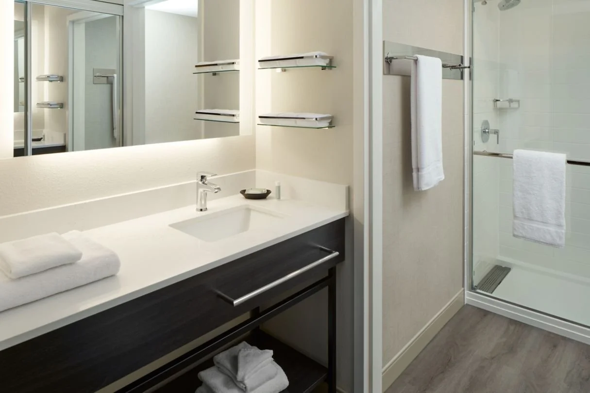 Hotel guestbath with wood and quartz vanity, large mirror and walk-in shower, all neutral finishes