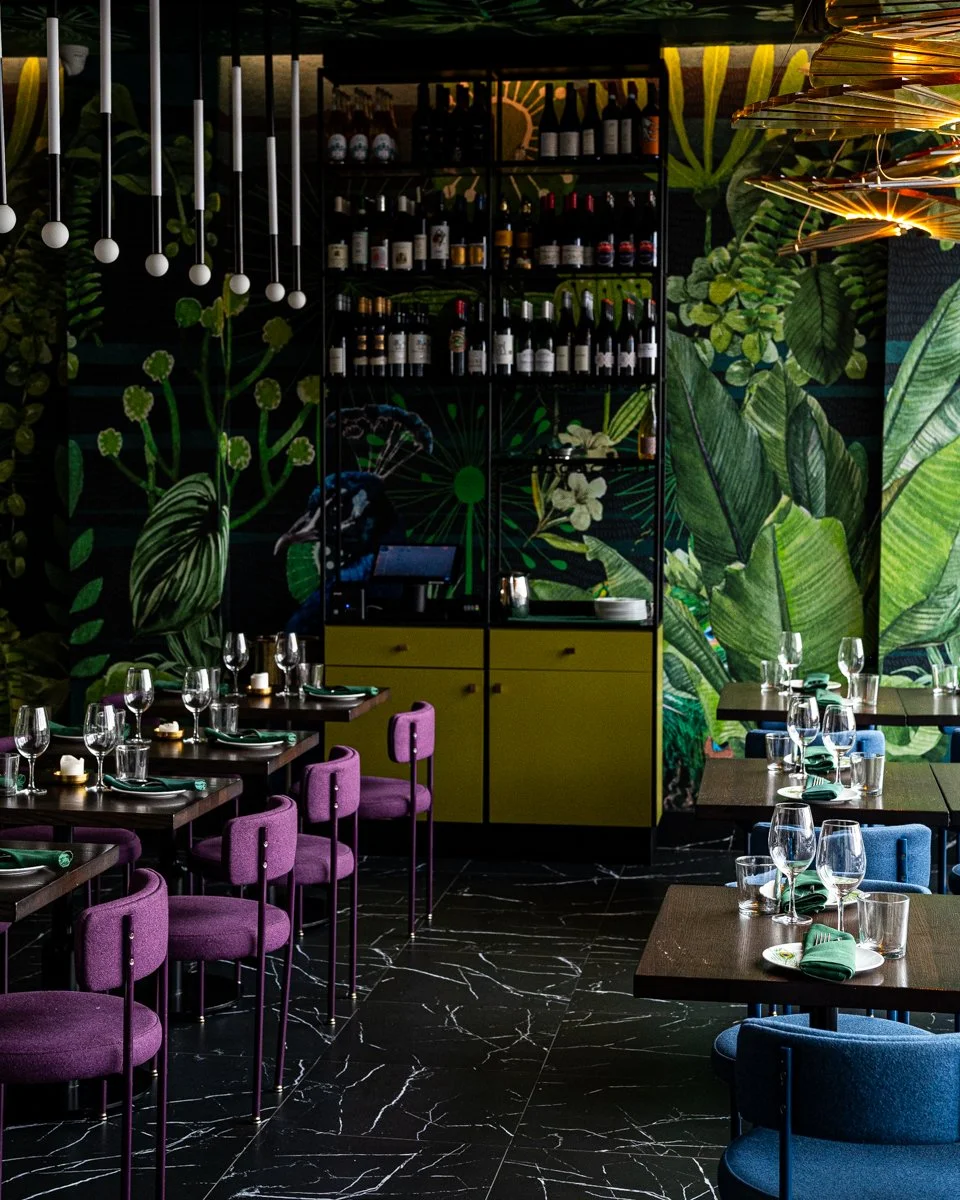 Restaurant interior with blue and purple chairs at wood tables with plant mural on walls, overhead glowing chandeliers and green service station