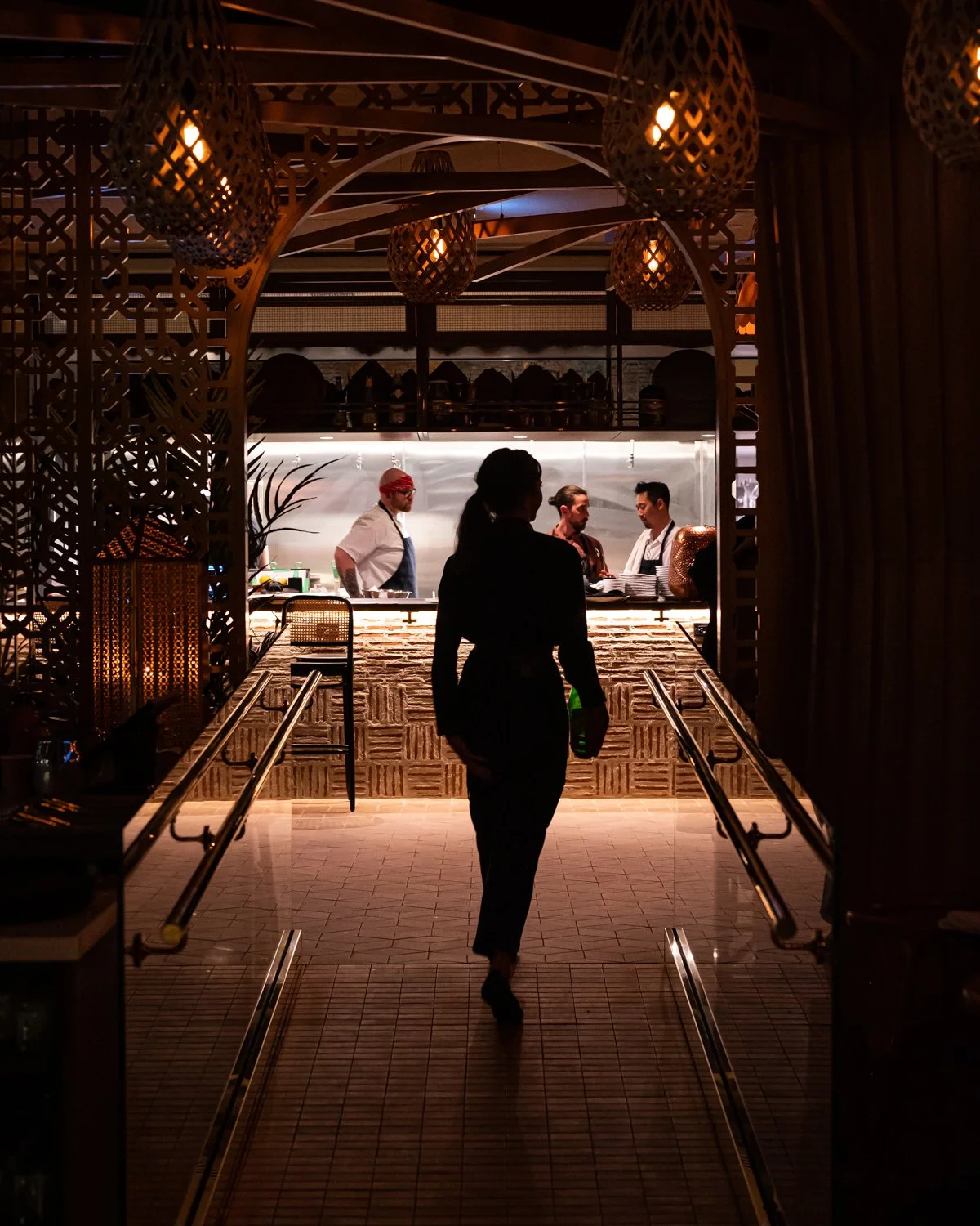 Evening setting of restaurant looking up ramp at brick bar through lattice screen with lantern lights overhead