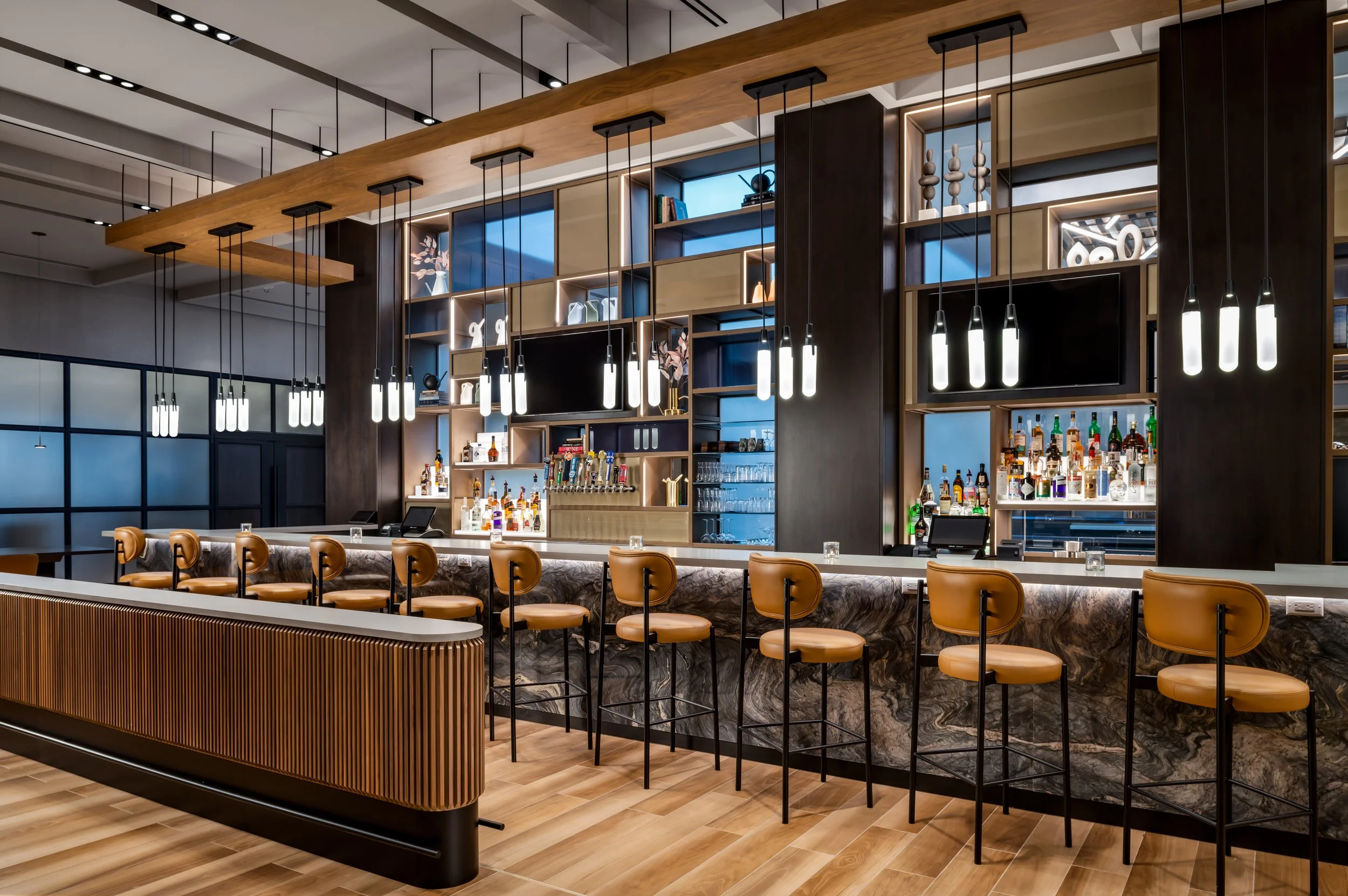 Hotel bar with veined stone surface, leather stools and suspended wood ceiling with trios of pendant lights