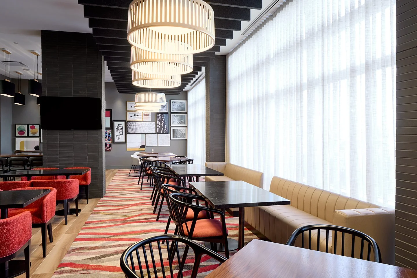 Hotel restaurant with red fabric seating, leather window banquettes, quartz top tables, and gallery wall art with overhead pendant lighting and grey stone walls