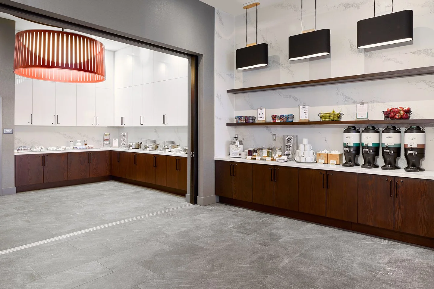 Buffet food service area with wood stained lower cabinets, quartz countertops, and white upper cabinets with accent black and red pendants