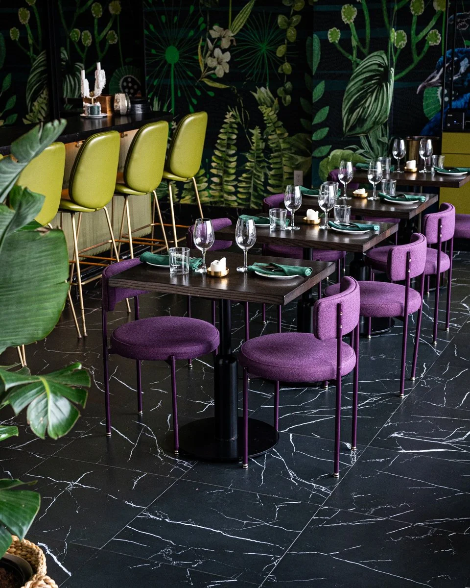 Restaurant interior with purple chairs at wood tables and green bar stools at green bar with green plant mural on walls and black marble floor tile