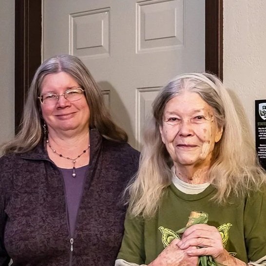 Two women standing indoors in front of a closed door, smiling, with the older woman holding a small item in her hands.