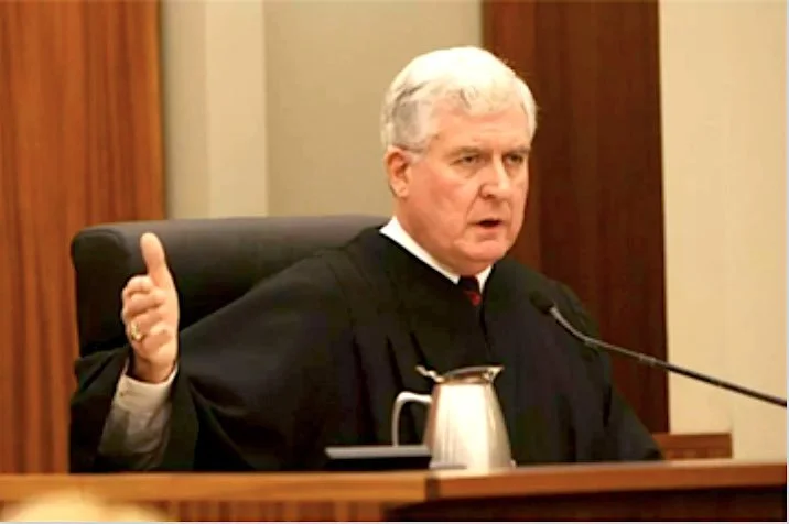 A judge in a black robe seated at a bench, gesturing with his hand, with a microphone and a pitcher of water in front of him.