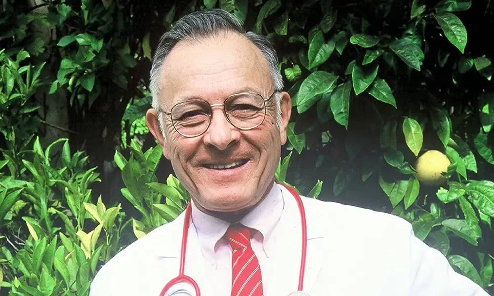 A smiling man in a white doctor's coat and red striped tie, wearing glasses and a stethoscope around his neck, standing outdoors in front of green leafy plants and a lemon on a tree.