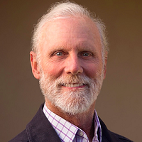 A close-up portrait of an older man with white hair and a beard, wearing a checkered shirt and a dark blazer, smiling at the camera.
