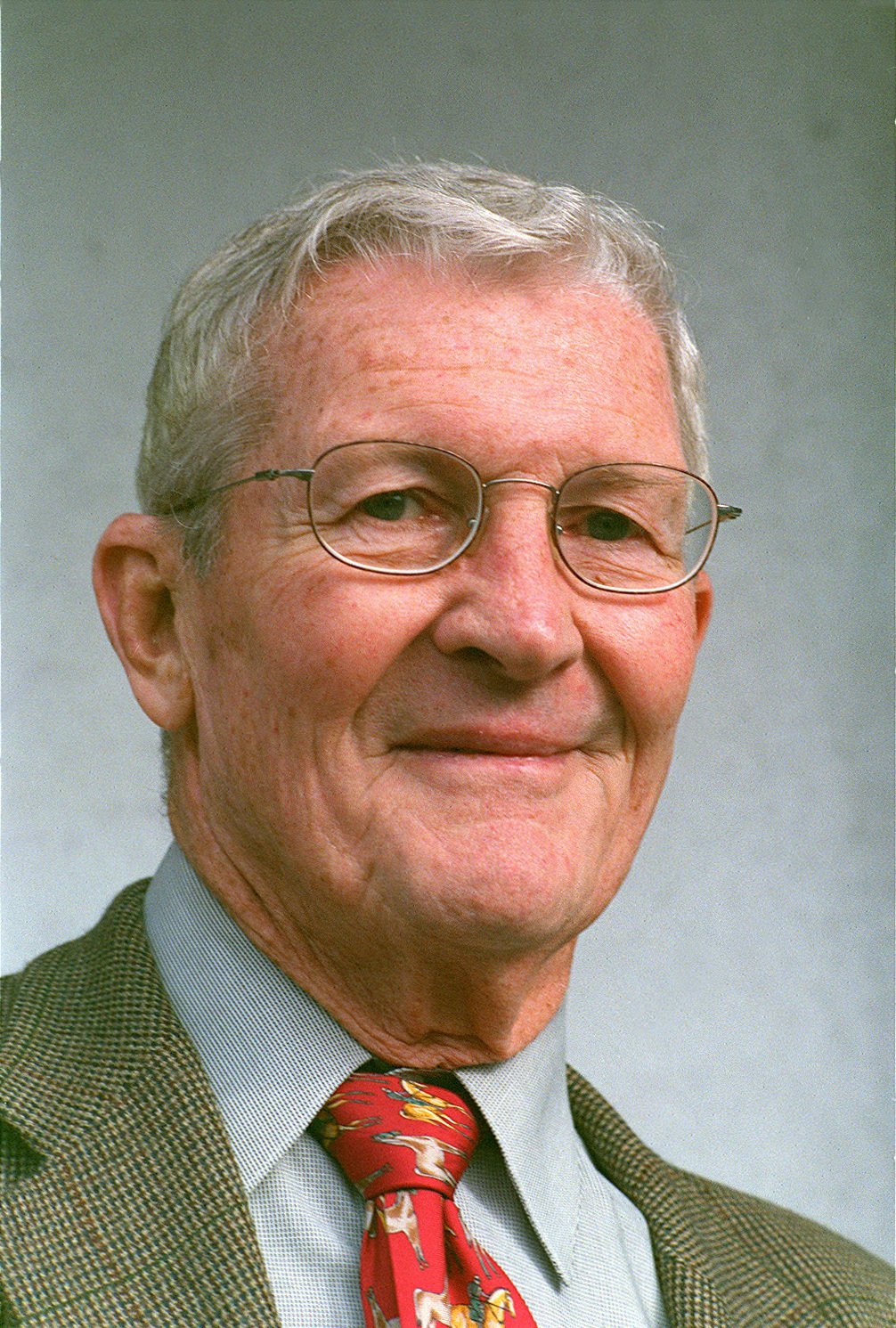 A close-up portrait of an elderly man with gray hair, wearing glasses, a white shirt, a patterned blazer, and a red tie with a horse pattern.