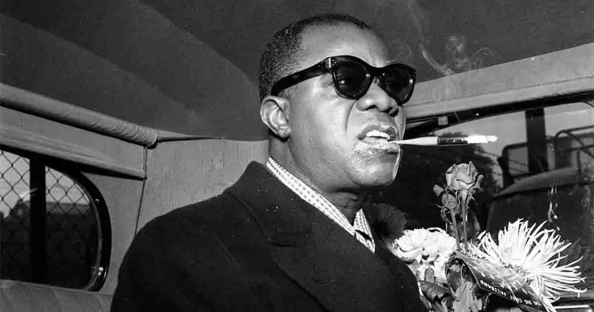 A man wearing sunglasses and a suit, holding a cigarette in his mouth, with flowers in his hand, inside a vehicle.