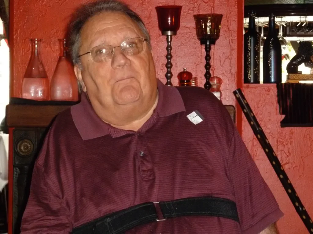 A middle-aged man with glasses and gray hair, wearing a maroon polo shirt with a black strap around his chest, sitting in a room with red textured walls and decorative vases and candlesticks on a shelf behind him.
