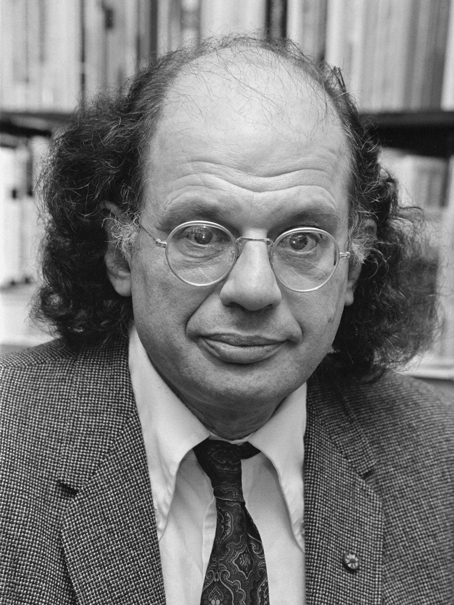 A black and white photo of a man with curly hair, glasses, and a mustache, wearing a checked blazer, white shirt, and patterned tie, sitting in front of bookshelves.