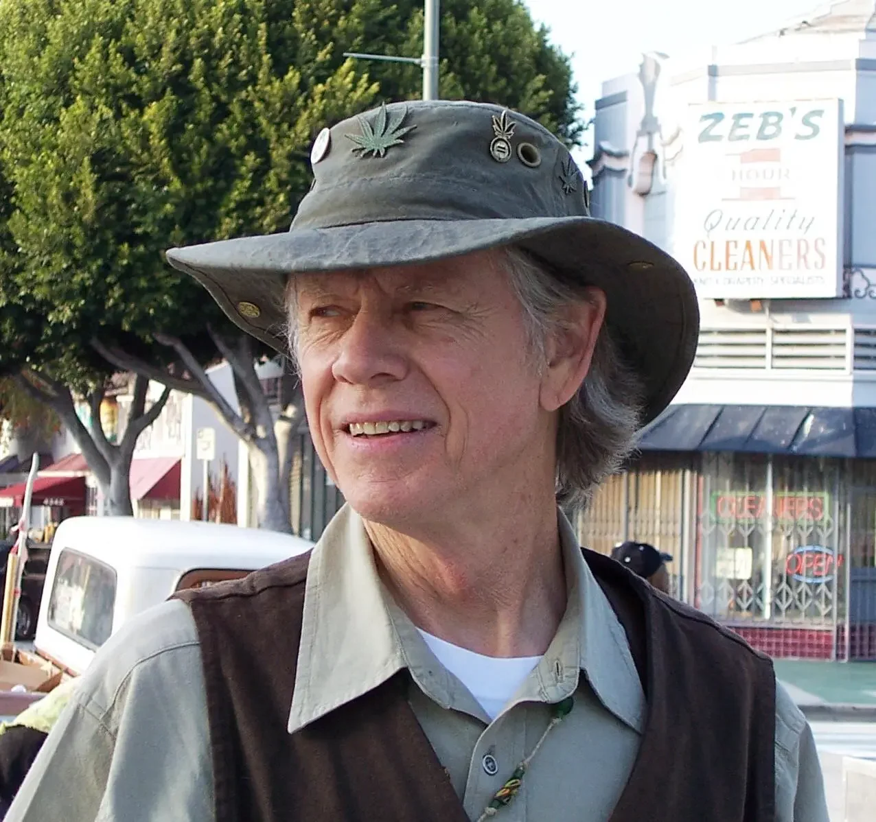 Smiling older man with gray hair wearing a beige hat decorated with marijuana leaf pins stands outdoors near a storefront with a sign that reads 'Zeb's'. Trees and parked cars are visible in the background.