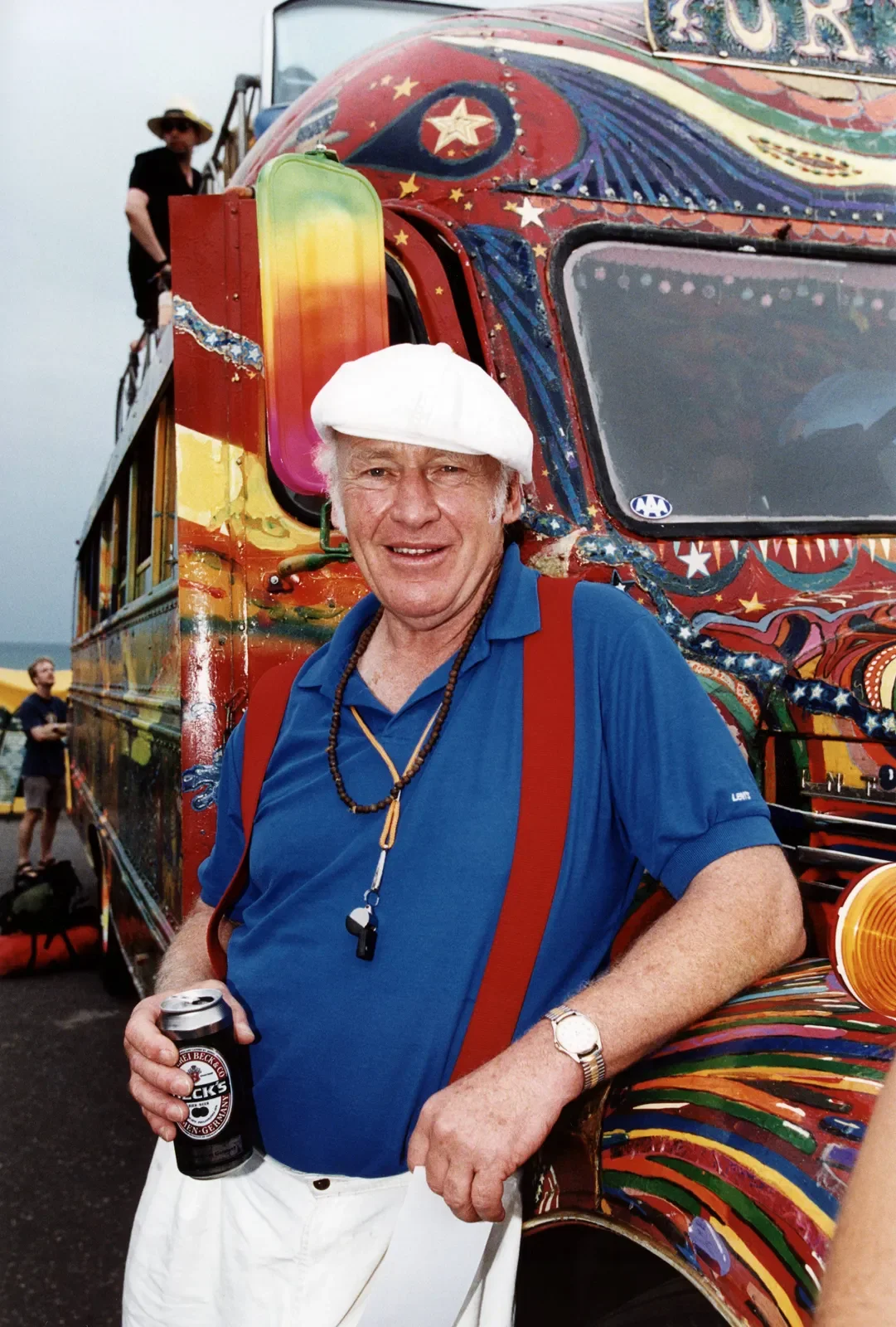 An older man with white hair wearing a white cap, blue shirt, red suspenders, and a watch, holding a can of beer, standing in front of a colorful, artistic vehicle with a painted elaborate design featuring stars and swirls. There are other people in the background.