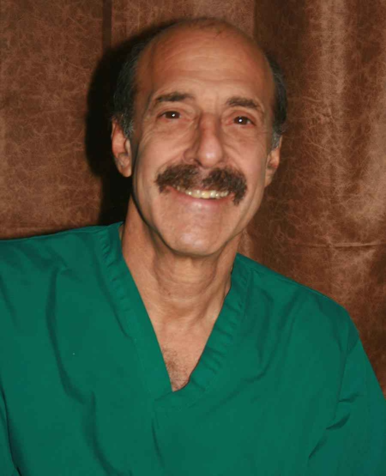A man with gray hair and a mustache, smiling, wearing green medical scrubs, posed against a brown textured background.