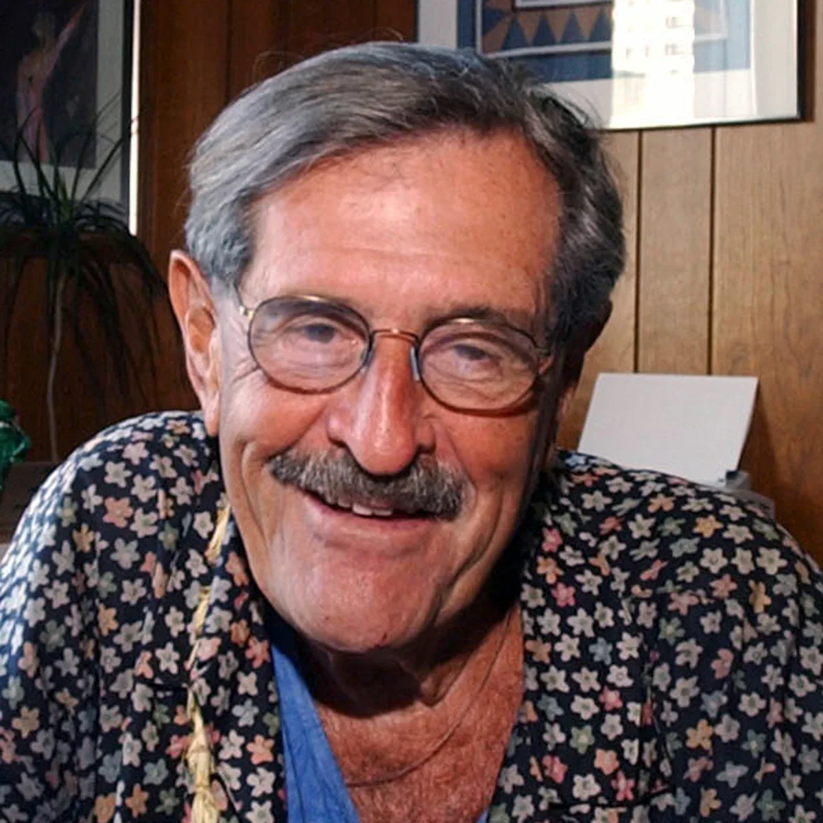 An elderly man with gray hair, glasses, and a mustache, smiling in a wooden-paneled room.