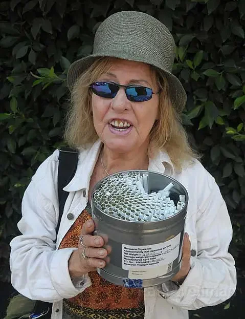An older woman wearing a gray hat, blue sunglasses, and a white jacket, holding a gray metal round container of cigarettes with a leafy green background.