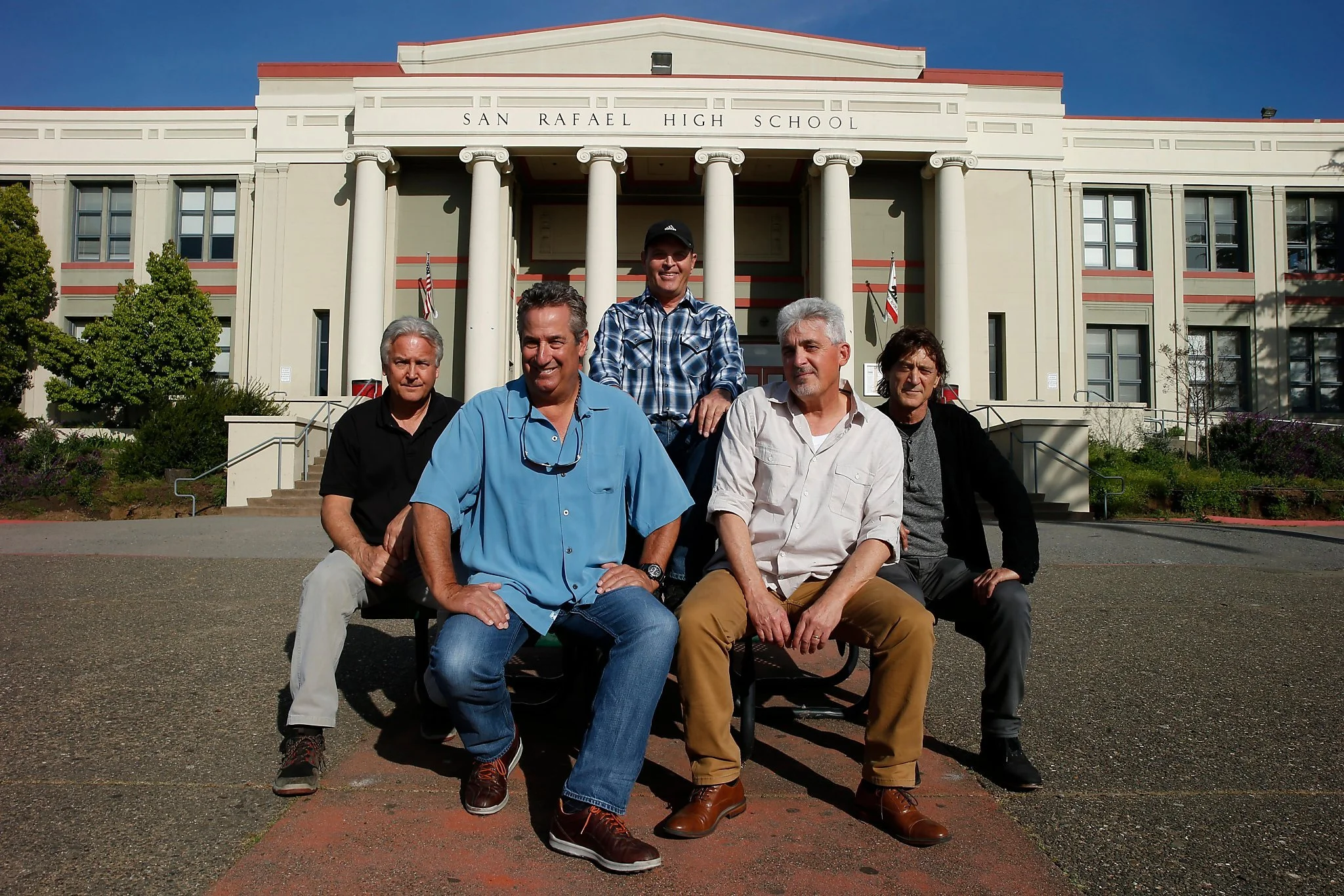 Group of five men sitting on a bench in front of a high school building labeled 'San Rafael High School'; clear sky and trees nearby.