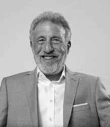 A smiling older man with gray hair and a beard, wearing a light-colored suit jacket and a white shirt, in black and white.