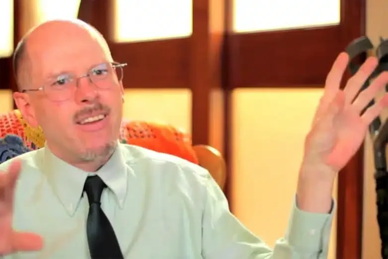A man with glasses and a bald head, wearing a white shirt and black tie, sitting in a room with wooden windows, gesturing with his hands.