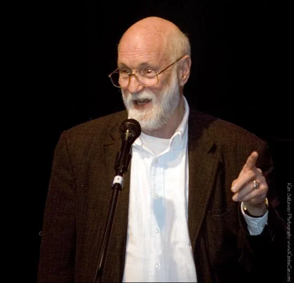 An elderly man with glasses and a beard, wearing a dark blazer and white shirt, speaking into a microphone during a presentation or speech.