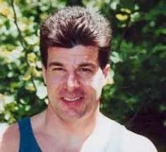 A young man with dark hair and a smile, wearing a tank top, outdoors with greenery in the background.