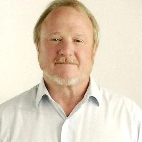 A middle-aged man with light hair and a beard, wearing a white collared shirt, smiling, against a neutral background.