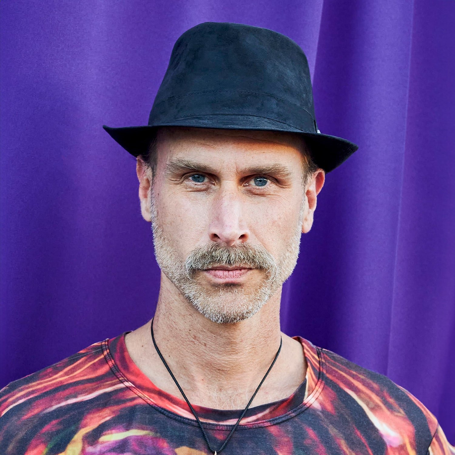 A man with short gray hair and a gray beard wears a black fedora hat, a colorful tie-dye shirt, and a black necklace, standing in front of a purple background.