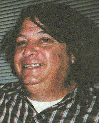 A smiling woman with dark hair and a side part, wearing a plaid shirt, indoors with horizontal blinds in the background.