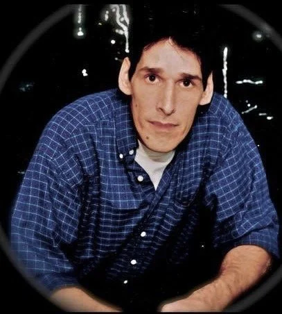 A man with dark hair and a serious expression wearing a blue checkered shirt, leaning forward in front of a dark background.