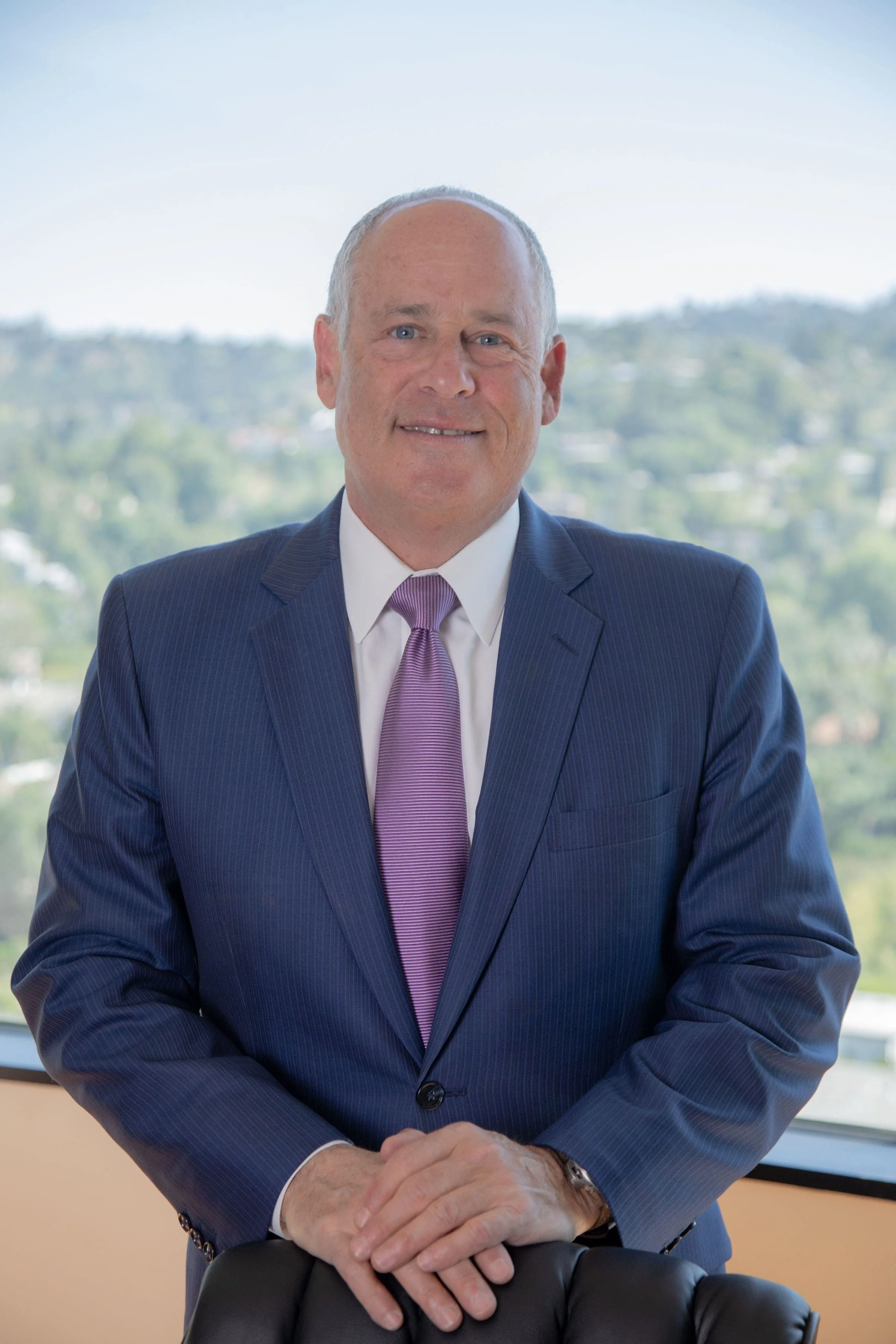 A middle-aged man in a dark blue suit, white shirt, and purple tie sitting by a window with a cityscape and green hills in the background, smiling at the camera.