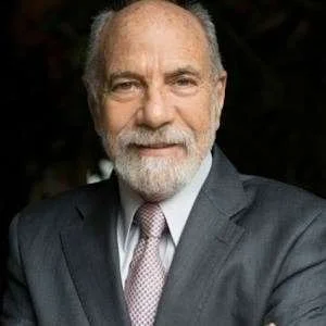 A portrait of an older man with white hair and a beard, wearing a suit and tie, against a dark background.