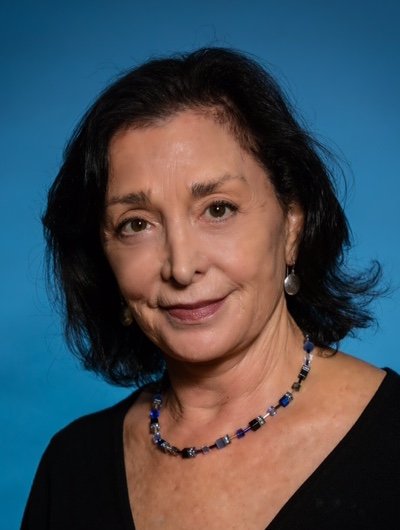 An elderly woman with black hair, wearing a black top, a beaded necklace, and earrings, against a blue background.