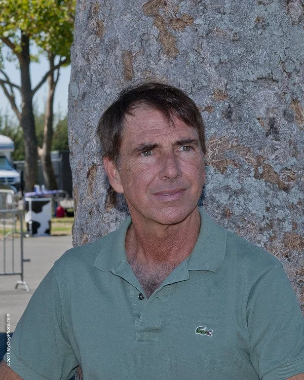 A middle-aged man with brown hair and a serious expression leaning against a tree trunk outdoors, wearing a green Lacoste polo shirt.