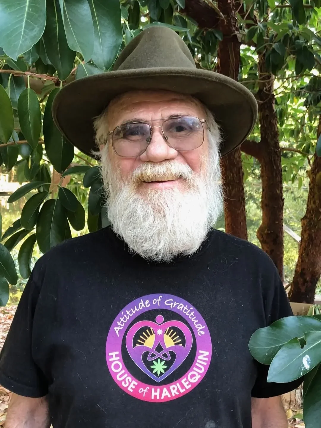 An elderly man with a long white beard and glasses wearing a wide-brimmed hat and a black t-shirt with a colorful logo reading 'Attitude of Gratitude House of Harlequin', standing outdoors among green leaves and trees.