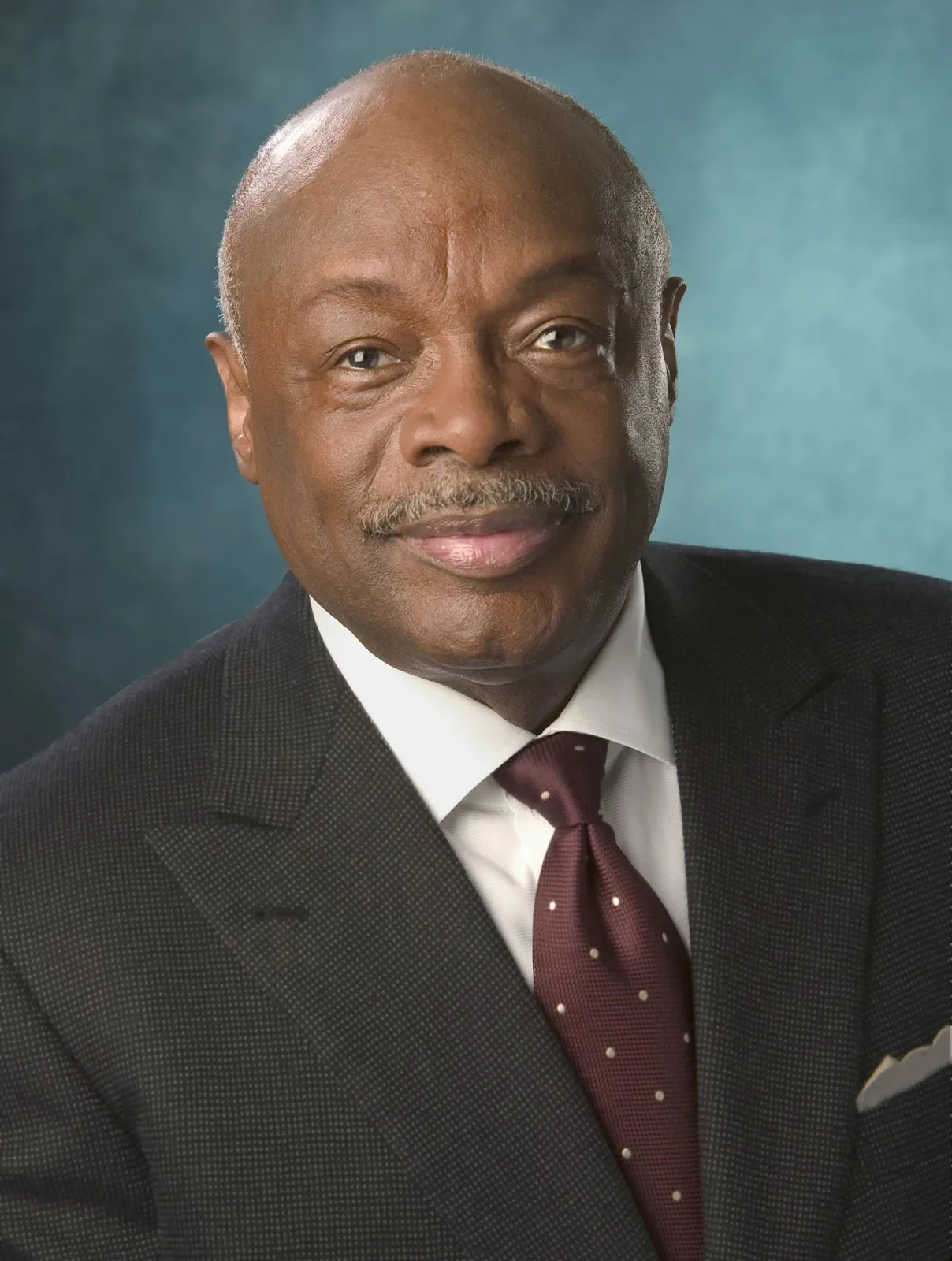 Headshot of an older man with a bald head, light mustache and goatee, wearing a dark suit, white dress shirt, and maroon tie with small polka dots against a blue background.
