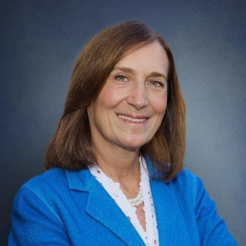 Portrait of a woman with shoulder-length brown hair, wearing a blue blazer and a white shirt with pearl necklace, smiling against a dark background.