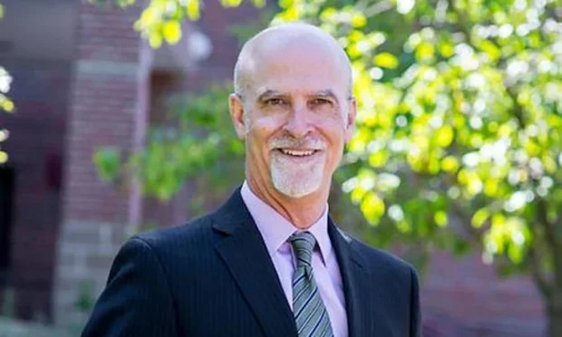 A smiling middle-aged man with a bald head and gray beard, wearing a dark suit, lavender shirt, and striped tie, standing outdoors in front of trees and a brick building.