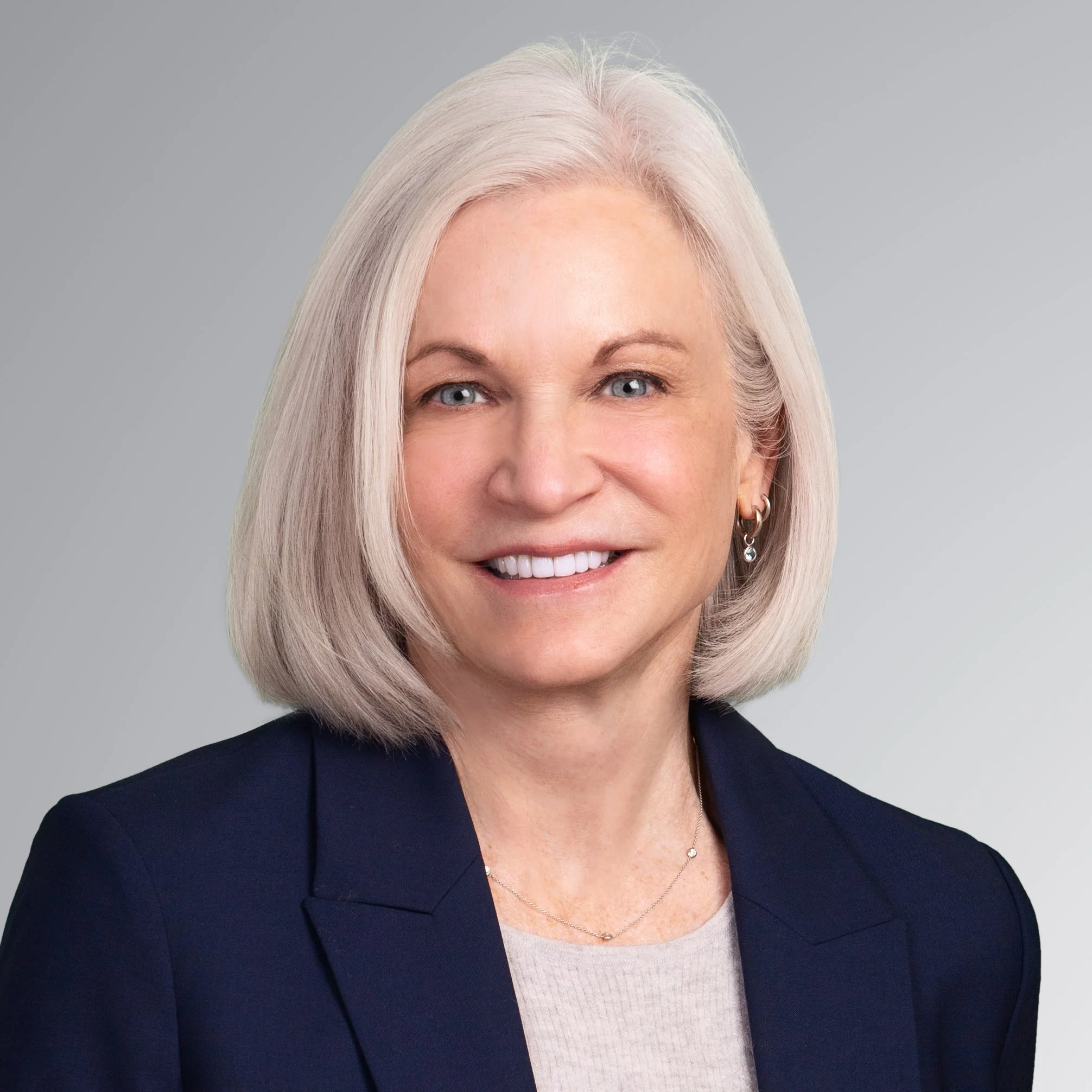 Close-up of a smiling older woman with blonde hair, wearing a navy blazer and a light-colored top, against a gray background.
