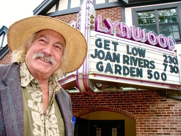 A man with long gray hair and a mustache wearing a sun hat and patterned shirt stands in front of a movie theater marquee that displays showtimes and titles, with a brick building and theater sign visible.