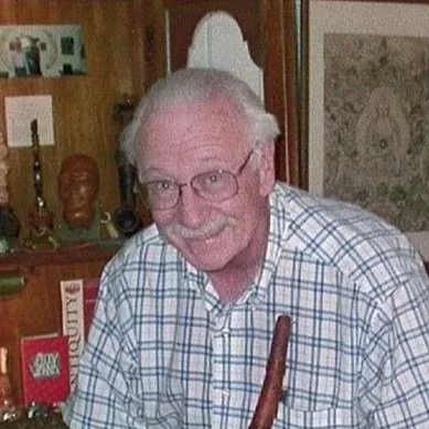 An elderly man with glasses and gray hair, smiling, wearing a checkered shirt, standing indoors with wooden decor and framed pictures behind him.