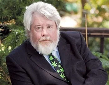A man with gray hair and a beard, wearing a suit and a tie with marijuana leaf patterns, sitting outdoors near greenery.