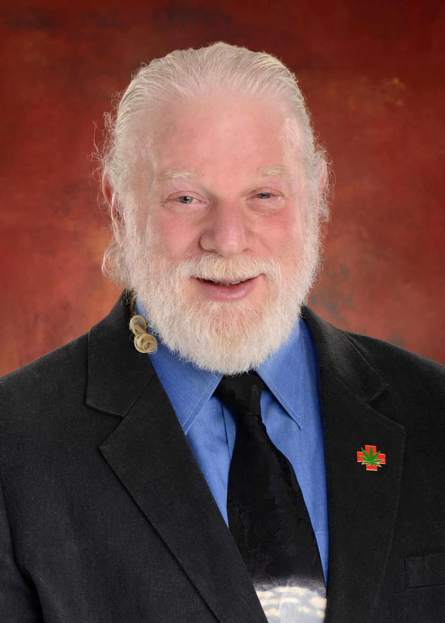 Close-up portrait of an elderly man with white hair, beard, and mustache, wearing a black suit, blue shirt, black tie, and a pin with a cannabis leaf on his lapel, against a brown textured background.