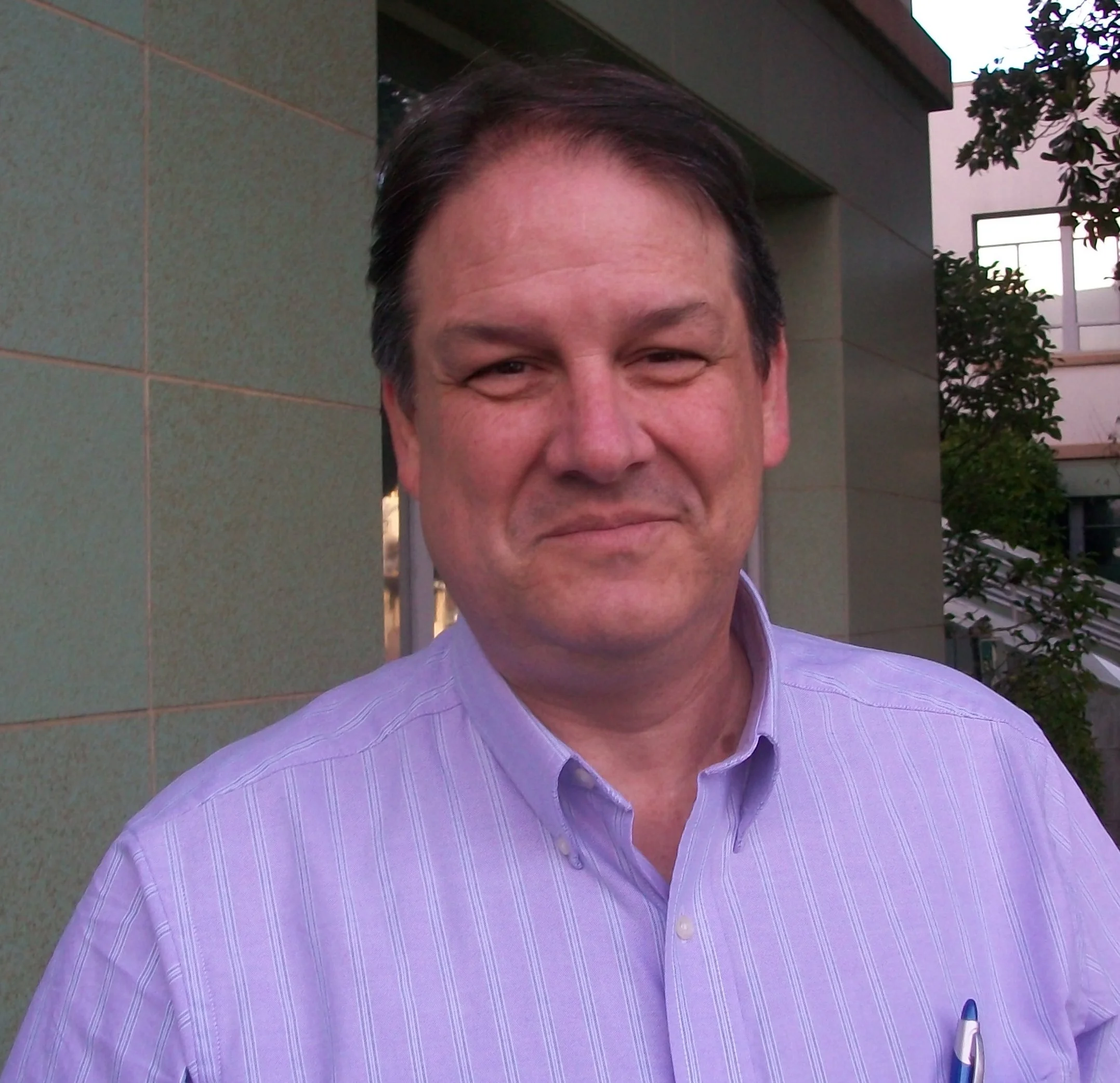 Close-up of a man smiling wearing a lavender striped dress shirt, standing outdoors near a building and some trees.