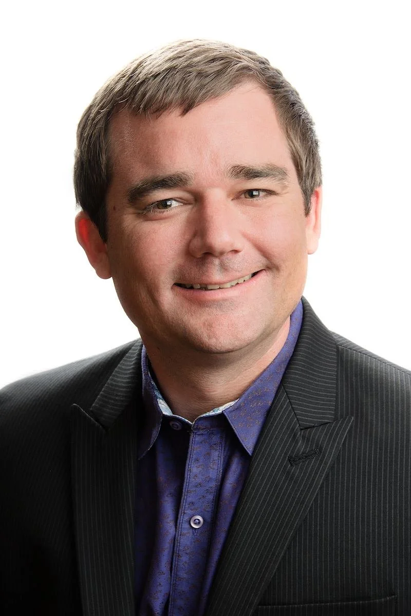 A professional portrait of a smiling man with short brown hair, wearing a dark pinstripe suit and a purple patterned dress shirt, against a white background.