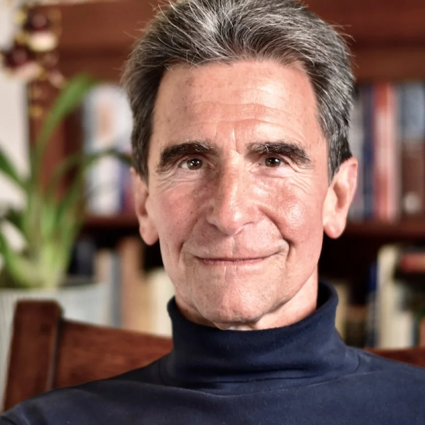 A middle-aged man with gray hair wearing a black turtleneck, smiling, sitting in a room with a bookshelf and a plant in the background.