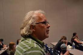 Side profile of an older man with white hair and glasses, sitting in a conference room filled with people.