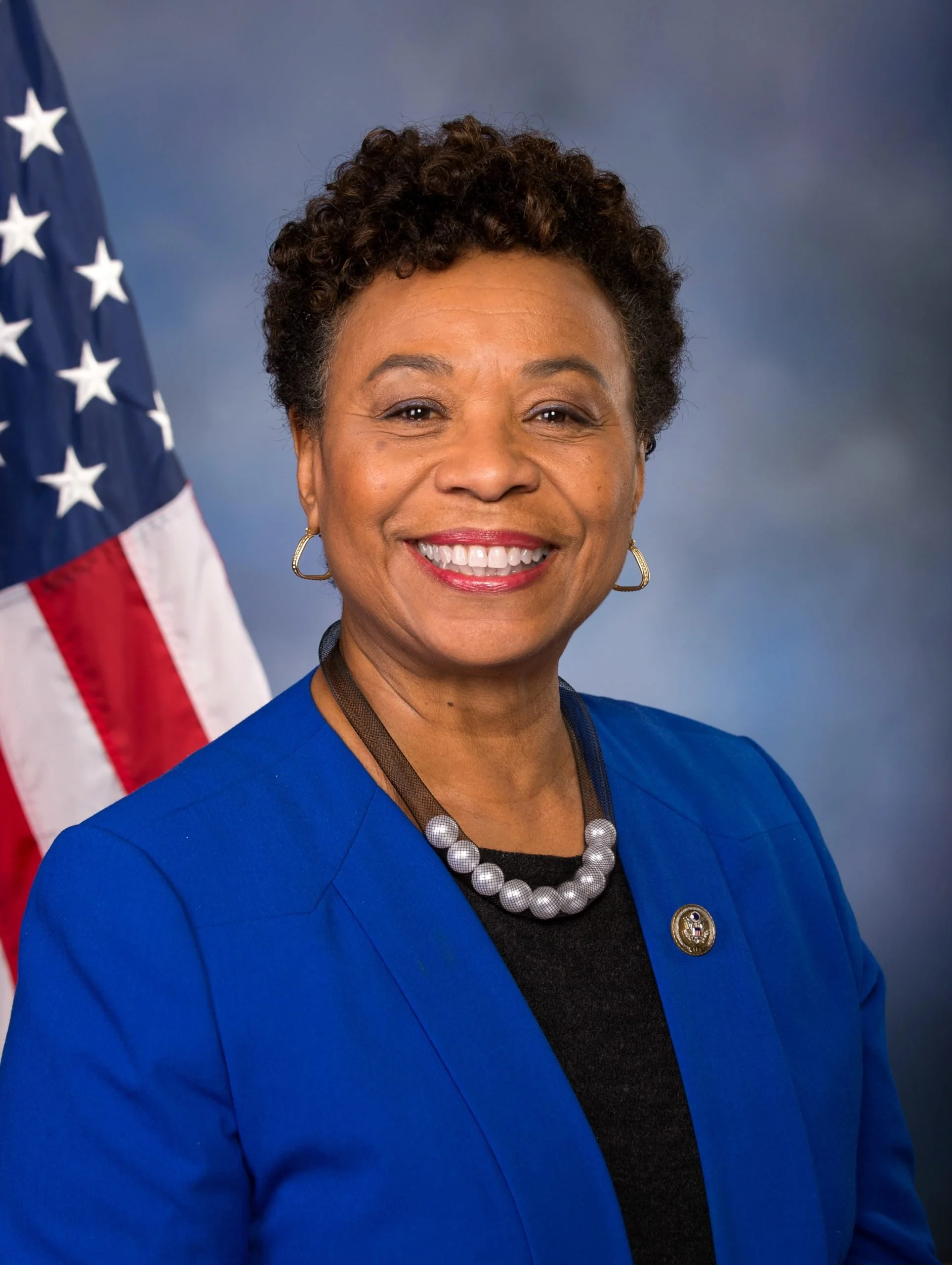 Close-up of an African American woman wearing a blue blazer, pearls, and a pin, smiling in front of a U.S. flag and a blue background.