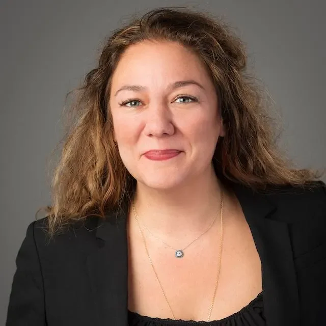 A woman with light skin and curly, shoulder-length brown hair, wearing a black blazer and a necklace with a small pendant, smiling slightly against a plain gray background.